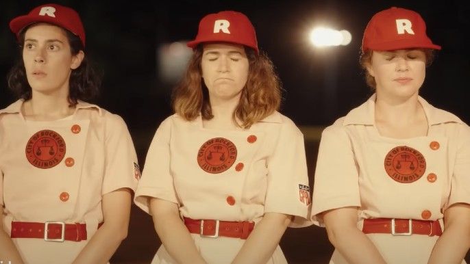 A League of Their Own show Abbi Jacobson as Carson and two other women squinting bright light in baseball field