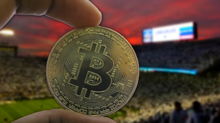 Image of Bitcoin physical gold coin being held by two fingers, against a football match background with a crowd.