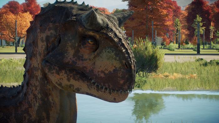 Jurassic World Evolution 2. The image is a close-up shot of a Carnotaurus dinosaur sitting on the left of the screen. The background is of the rest of its enclosure and some water. 