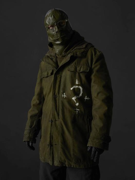 The Riddler stands wearing a khaki, green trench coat and a mask with glasses.