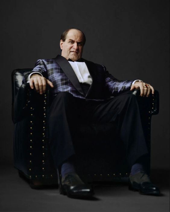 The Penguin sits down on a leather chair, wearing a checkered, blue suit.