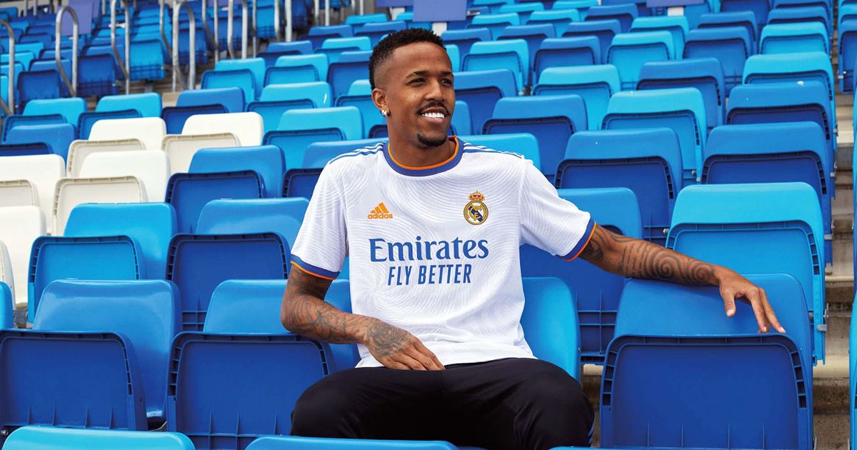 Éder Militão wearing a white Real Madrid kit featuring blue and orange branding while sat on a blue seat inside a stadium.