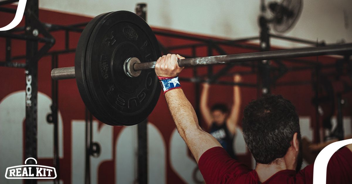 Someone in a red top and blue wrist wraps with a white star lifting a barbell with black weight plates overhead.