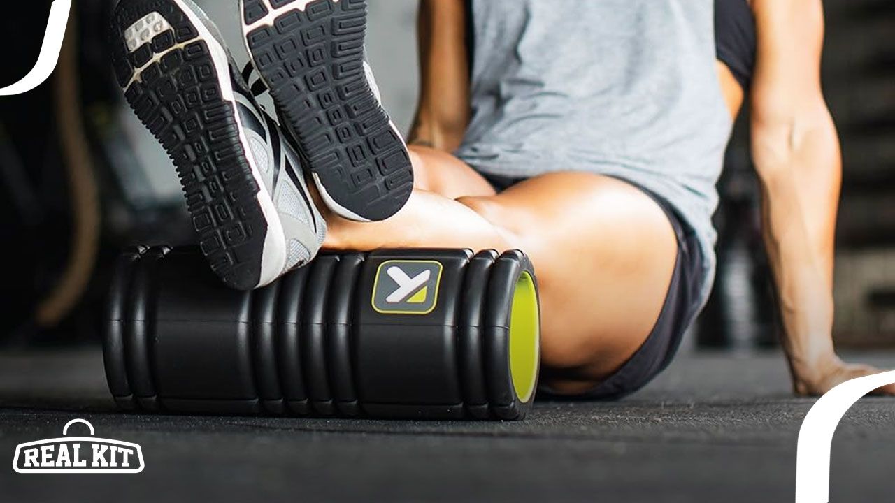 Someone in a grey vest and black shorts leaning their legs on a black foam roller with a green interior.