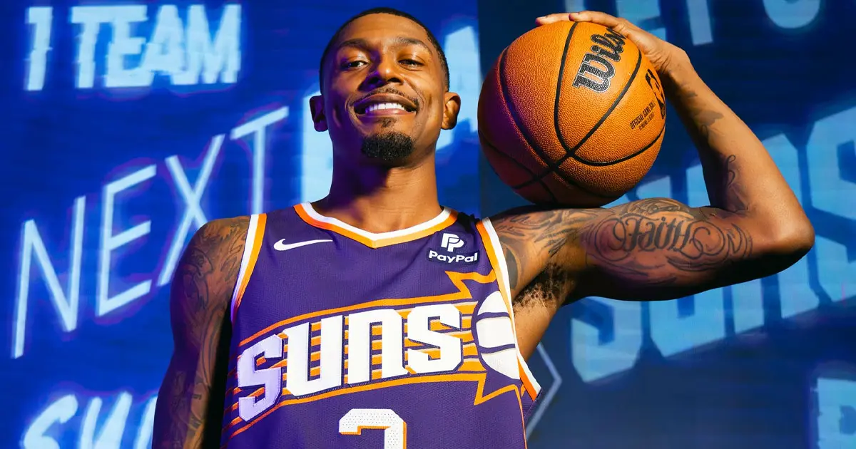 An NBA player holding an orange basketball wearing a purple jersey with white and orange trim.