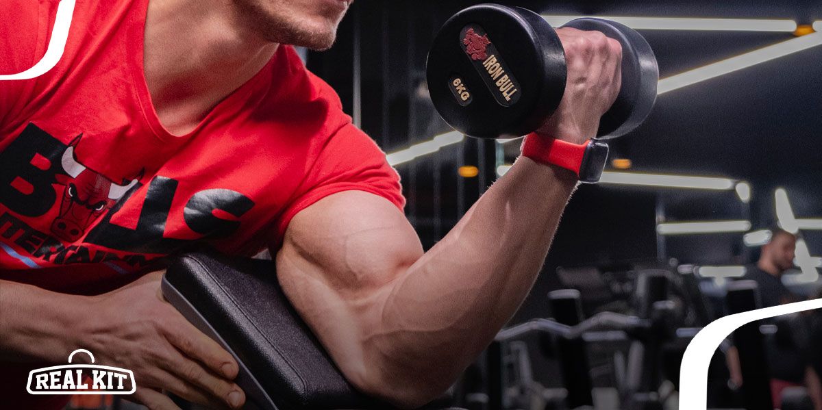Someone in a red Chicago Bulls t-shirt bicep curling a black 6kg dumbbell resting against a padded stand.