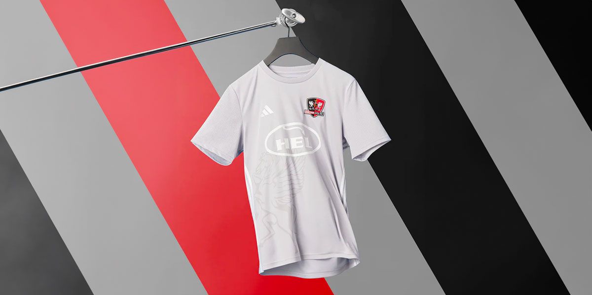 Exeter City adidas Away Kit product image of a light grey shirt featuring white branding and sponsors hanging in front of a grey, red, and black striped background.