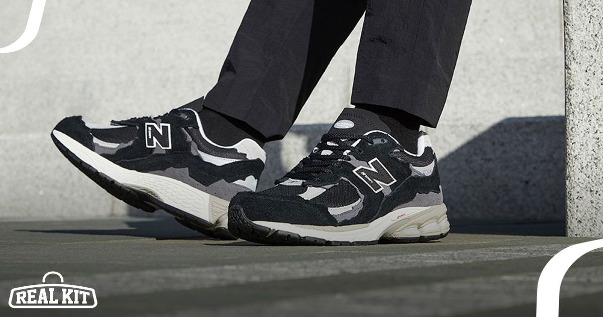 Someone in black trousers and socks wearing a pair of black New Balance shoes featuring an off-white midsole and grey details.