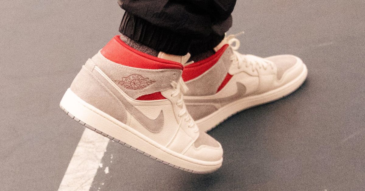 Someone in black cuffed trousers wearing a pair of light cream and brown Jordan 1 Mids featuring red trim.