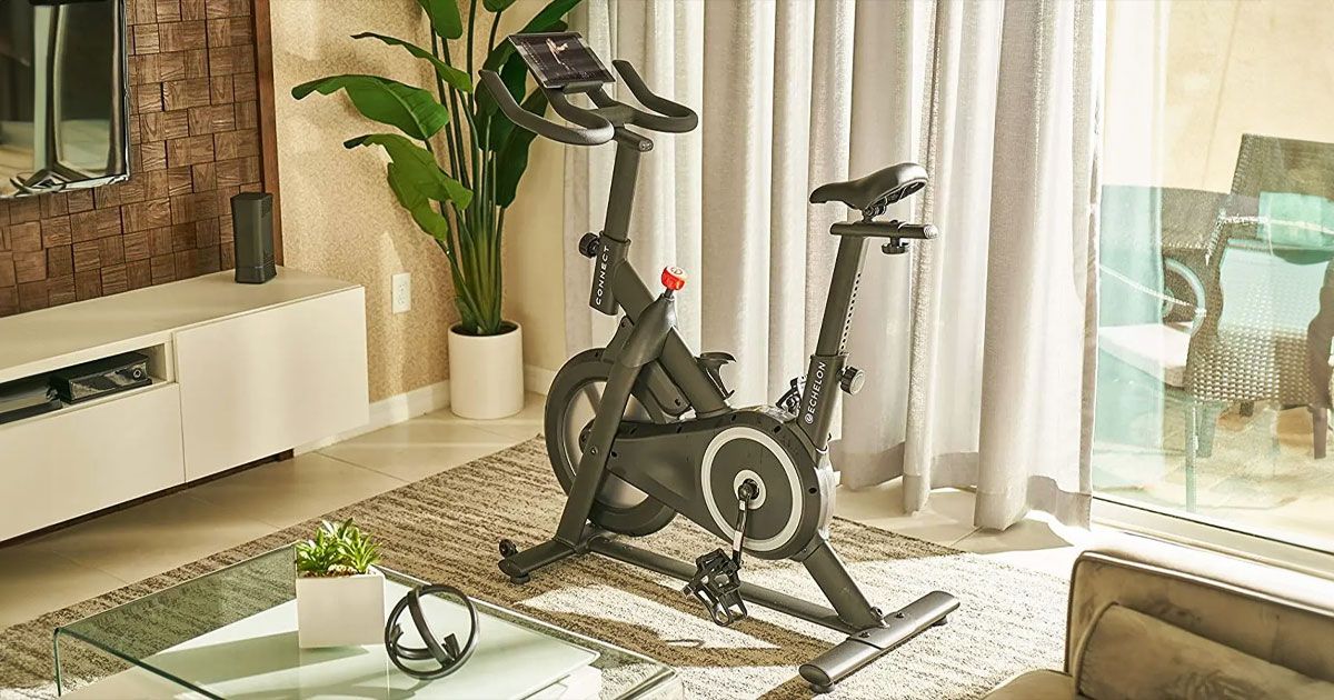 A black spin bike sat in a living room in front of a wall-mounted TV and next to a window.