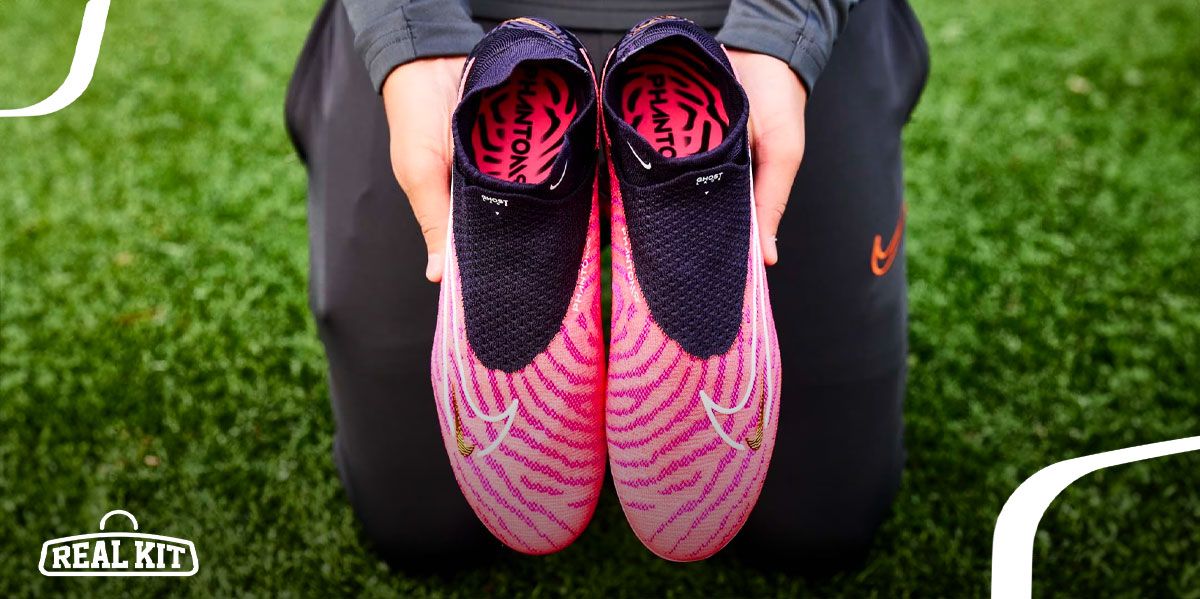Image of someone wearing black tracksuits holding a pair of pink and black laceless Nike football boots.