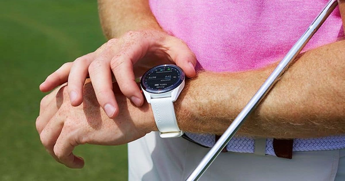 Someone in a pink shirt and white trousers with a golf club under their arm adjusting their white and black smartwatch.