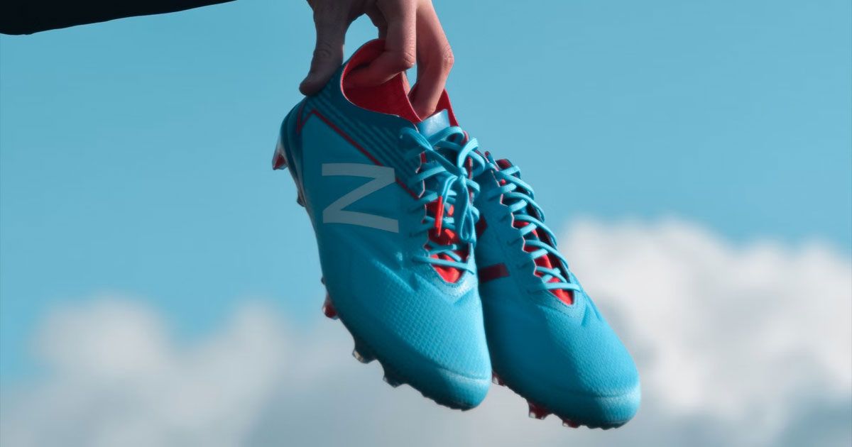 Someone holding up a pair of blue New Balance football boots with white branding on the side and red interiors.