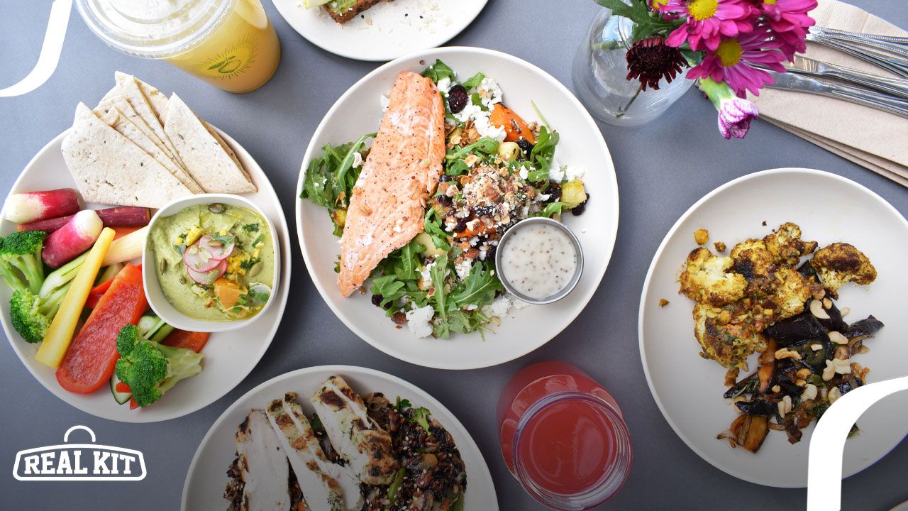 A plate of salmon and salad in the centre of the image, surrounded by other meals including pitta, fruit, chicken, and juice.