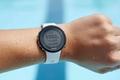 Image of a Garmin Swim 2 watch with a white strap on a wrist of someone standing by a pool.