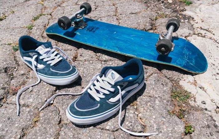 Vans product image of a pair of blue, unlaced sneakers next to a blue skateboard.