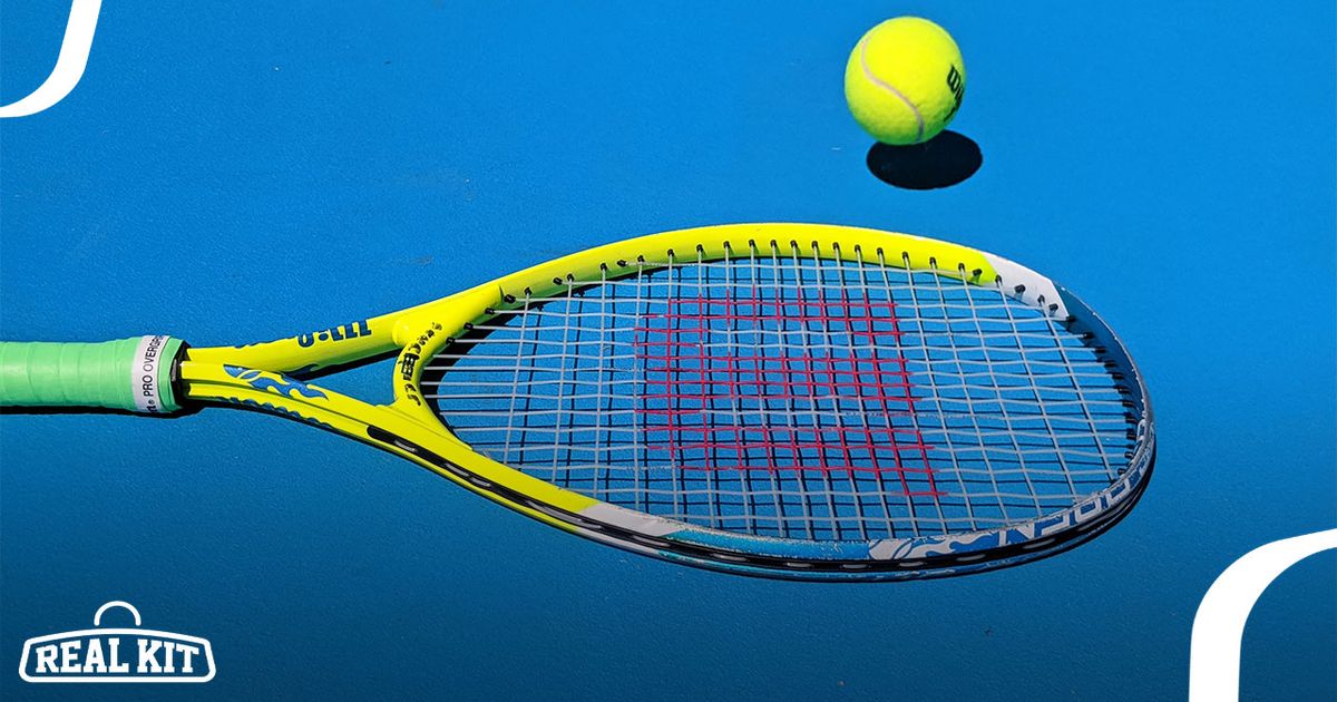 A yellow Wilson tennis racquet with a green handle laying on a blue court next to a yellow tennis ball.