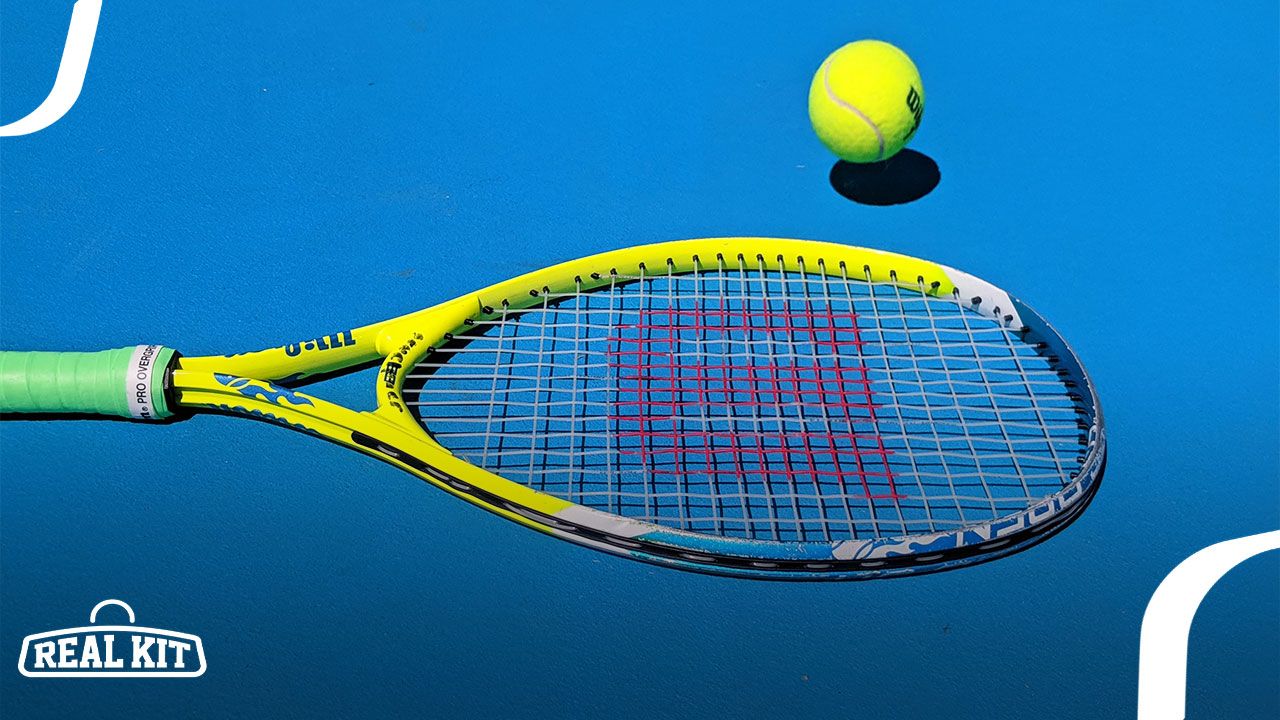 A yellow Wilson tennis racquet with a green handle laying on a blue court next to a yellow tennis ball.