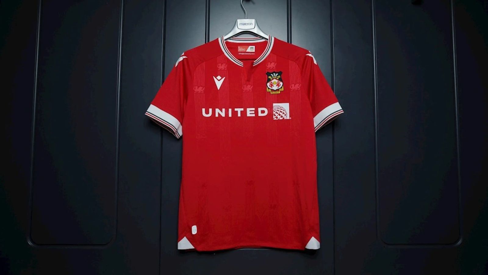 Wrexham Macron Home Kit product image of a red jersey featuring a white collar, sleeves, branding, and sponsorship in front of a black panelled background.