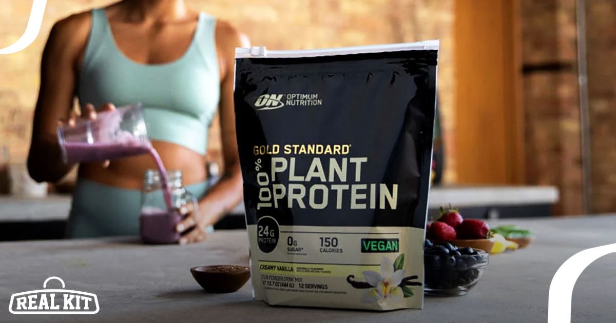 Image of a black package of plant protein featuring cream and gold branding, sat in front of someone wearing blue making a pink smoothie.