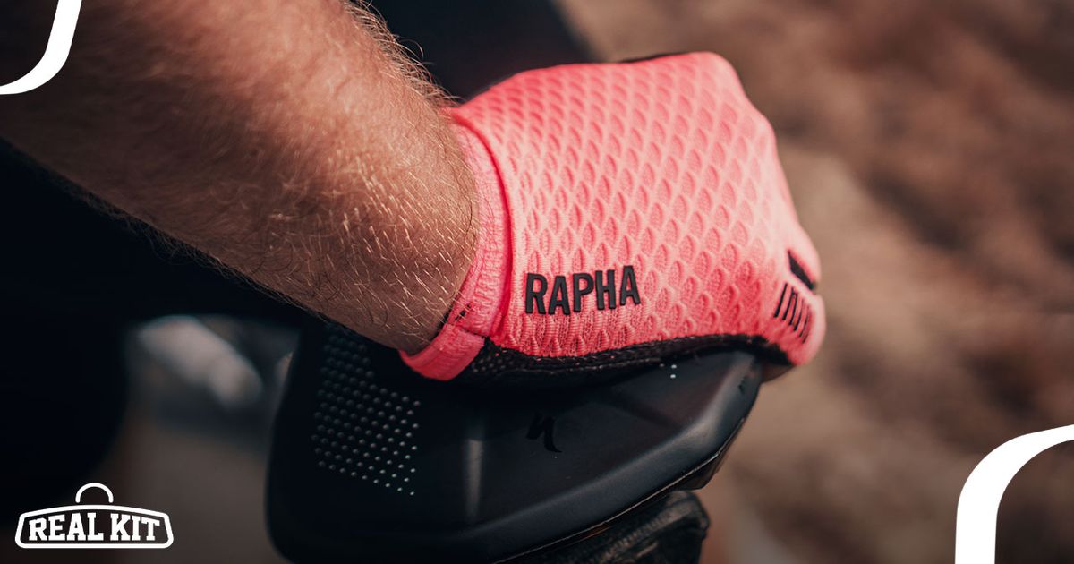 Close-up of someone wearing a bright pink glove with their hand on a black saddle, the glove featruing "RAPHA" in black down the side.