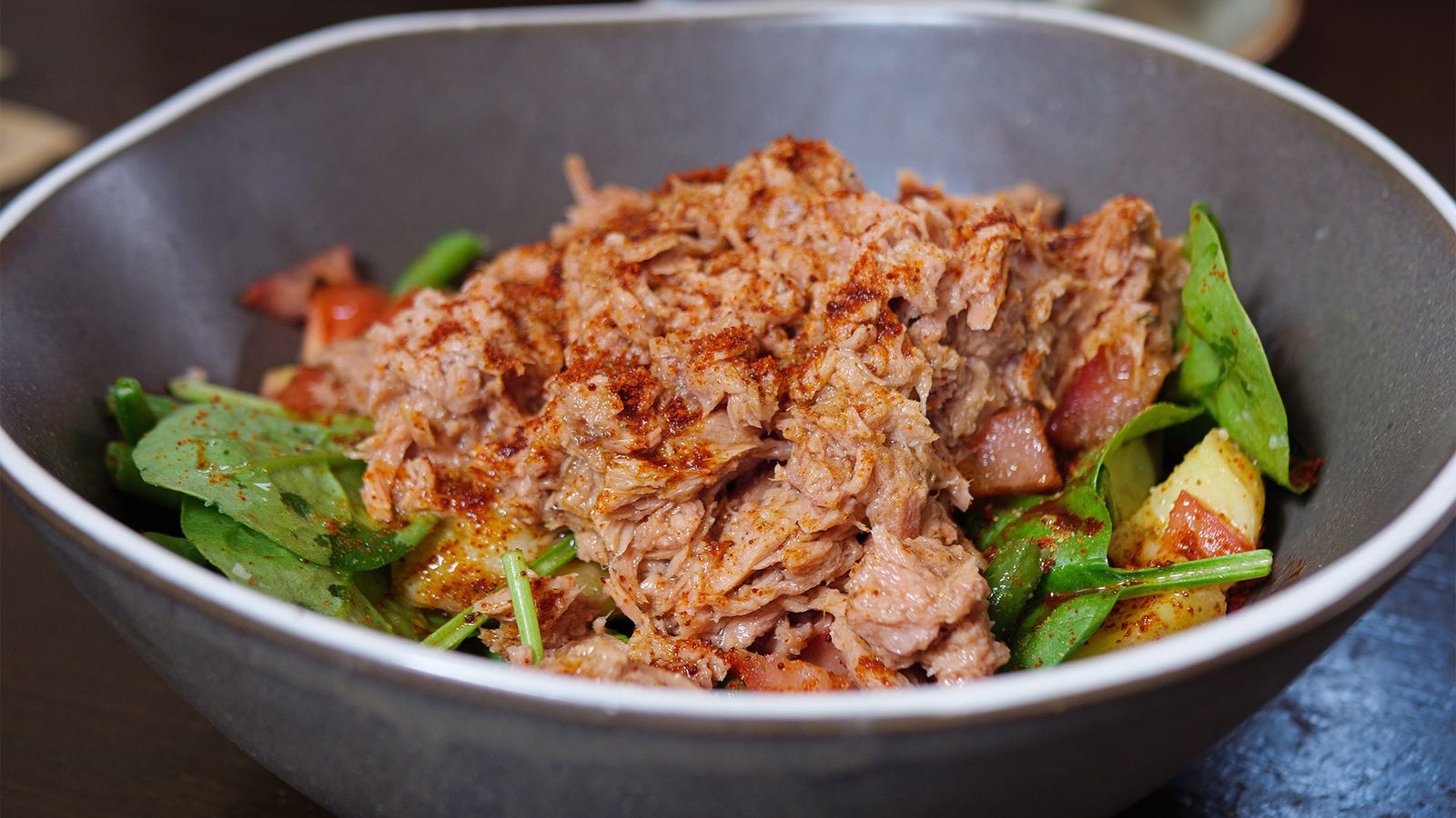 Close-up photo of a tuna salad with potatoes, tomatoes, spinach, and green beans sprinkled with smoked paprika seasoning and served in a grey bowl.