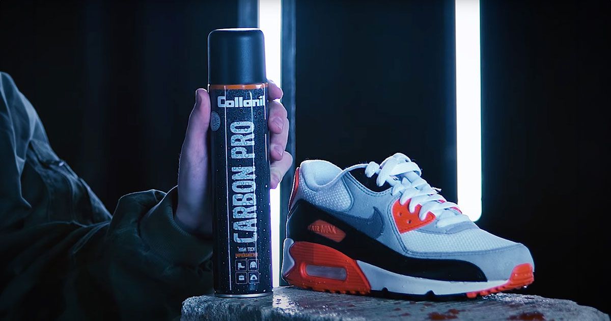 Someone in a black jumper holding a black can of sneaker protector spray in front of a white, black, and orange Nike Air Max.