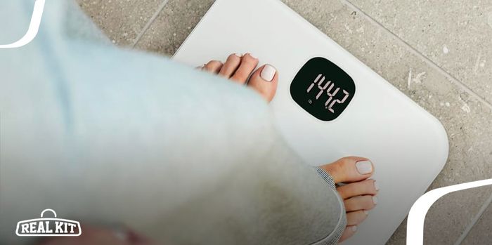 Close-up of someone in white trousers standing on a white Fitbit smart scale with a black digital display.