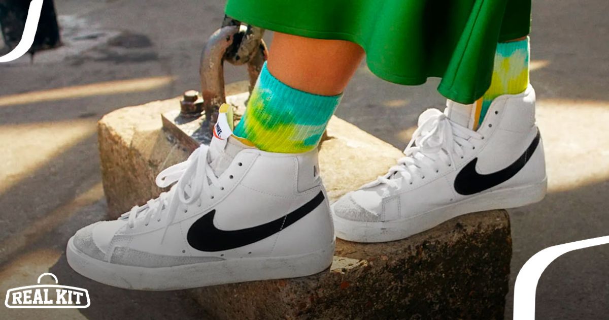 Someone in a green skirt and turquoise and yellow socks wearing white Nike Blazer Mids featuring black Swooshes.