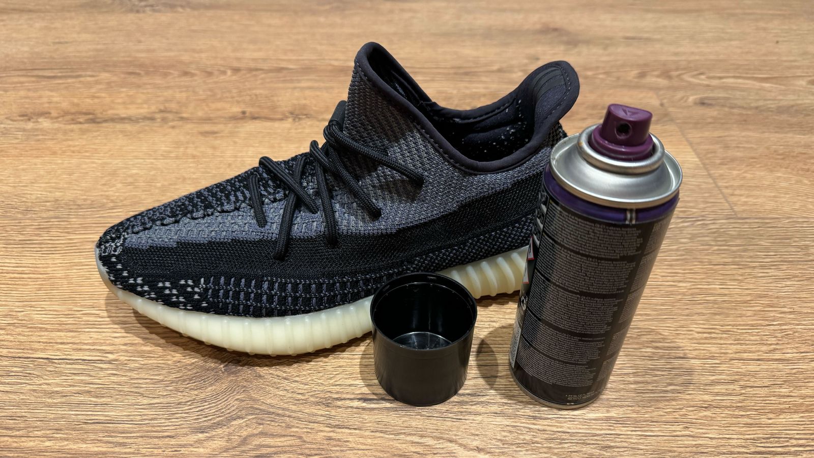 A black Crep Protect spray can with a purple spray nozzle next to a black knitted Yeezy with a gum sole.