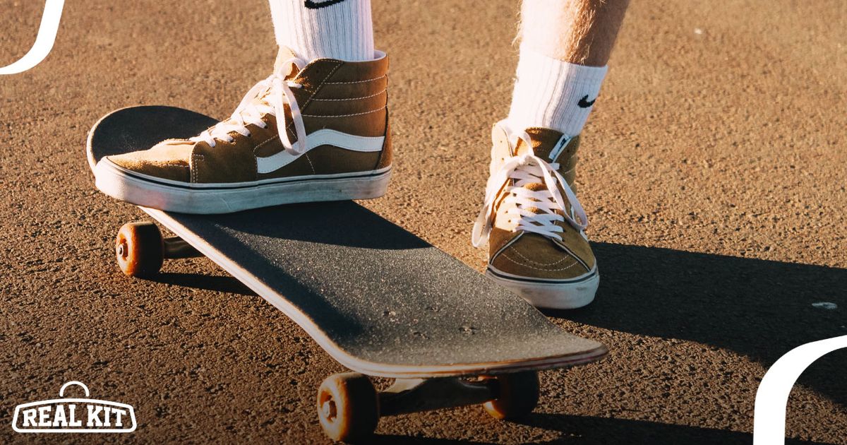 Person in white Nike socks wearing brown and white Vans Sk8 Hi sneakers standing on a skateboard.