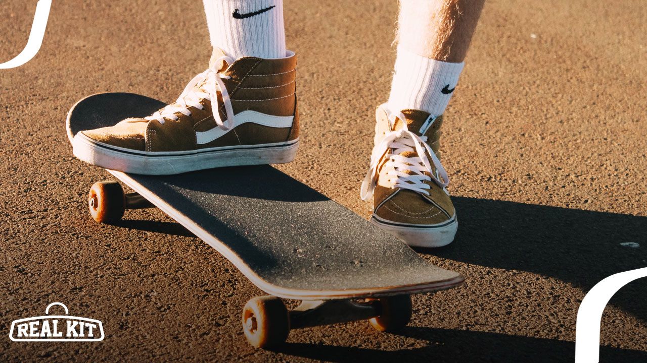 Person in white Nike socks wearing brown and white Vans Sk8 Hi sneakers standing on a skateboard.