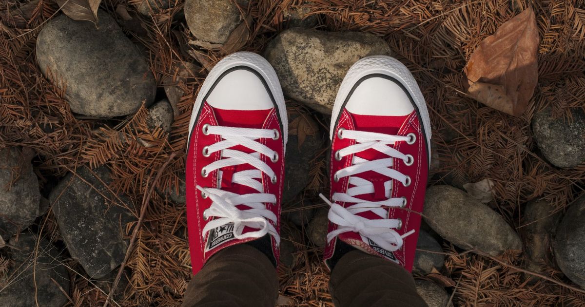 Someone standing on brown fallen leaves and rocks wearing a pair of red and white Converse Lows.
