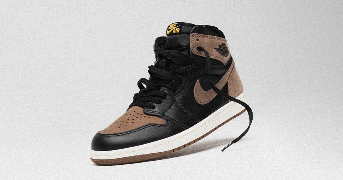 A black leather and brown suede Jordan 1 High with a white midsole and yellow branding on the tongue propped up.