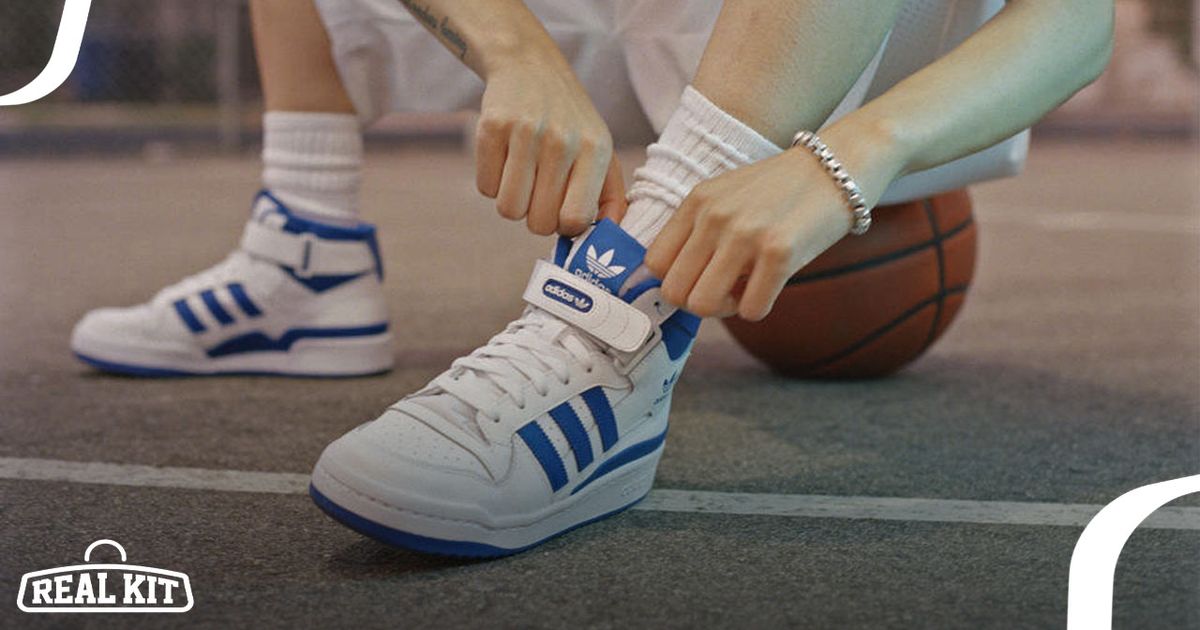 Image of a pair of bright white and blue adidas Forum sneakers on feet of someone sitting on a basketball.