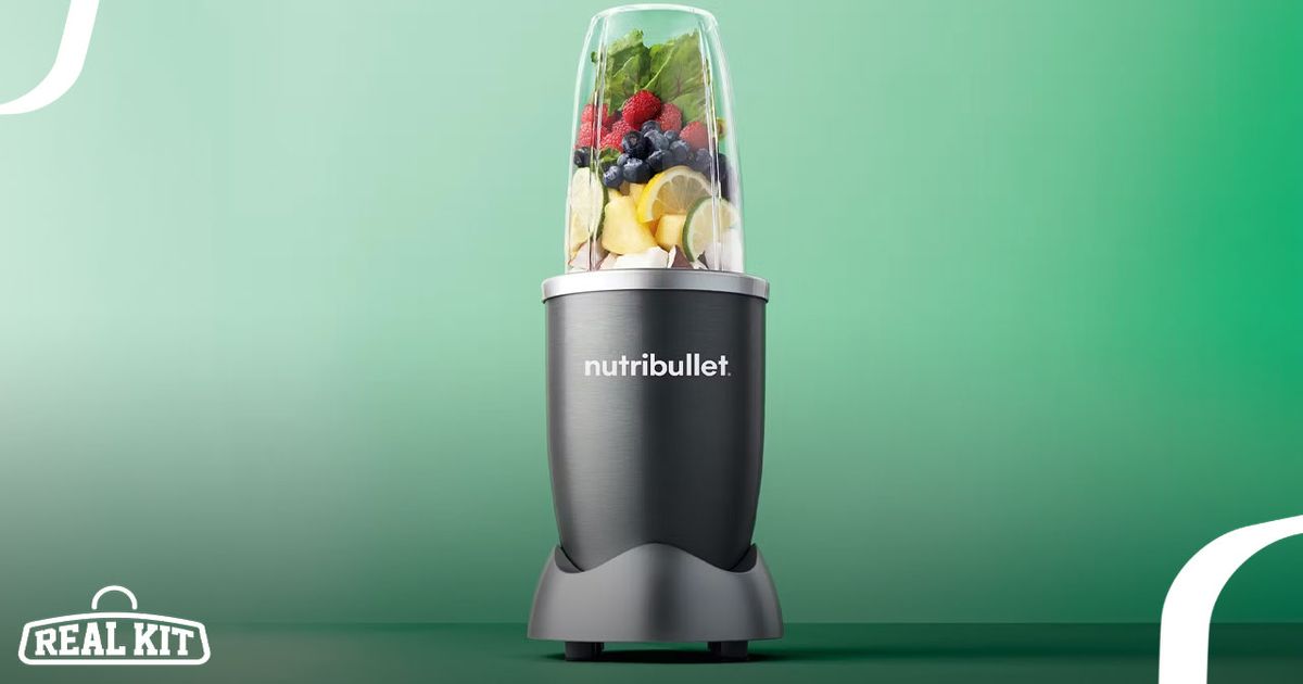 A grey blender with fruit and veg in the container above in front of a green backdrop.