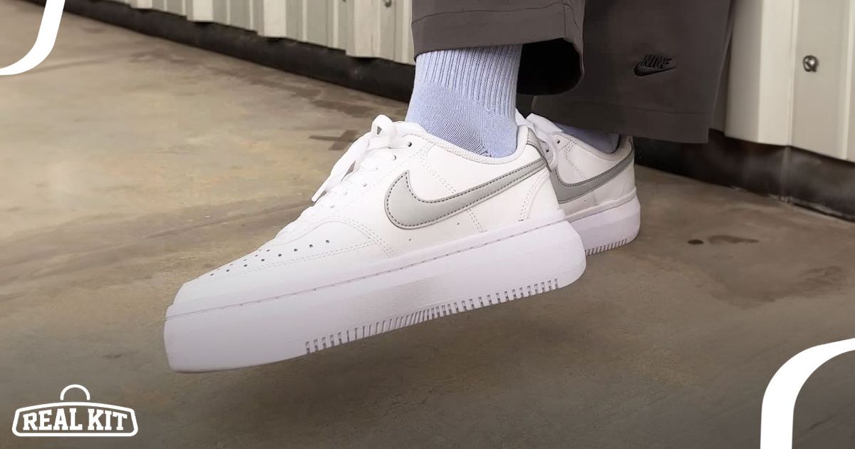 Someone in brown Nike trousers and light blue socks wearing a pair of white Nike Court Vision low-tops featuring grey Swooshes.