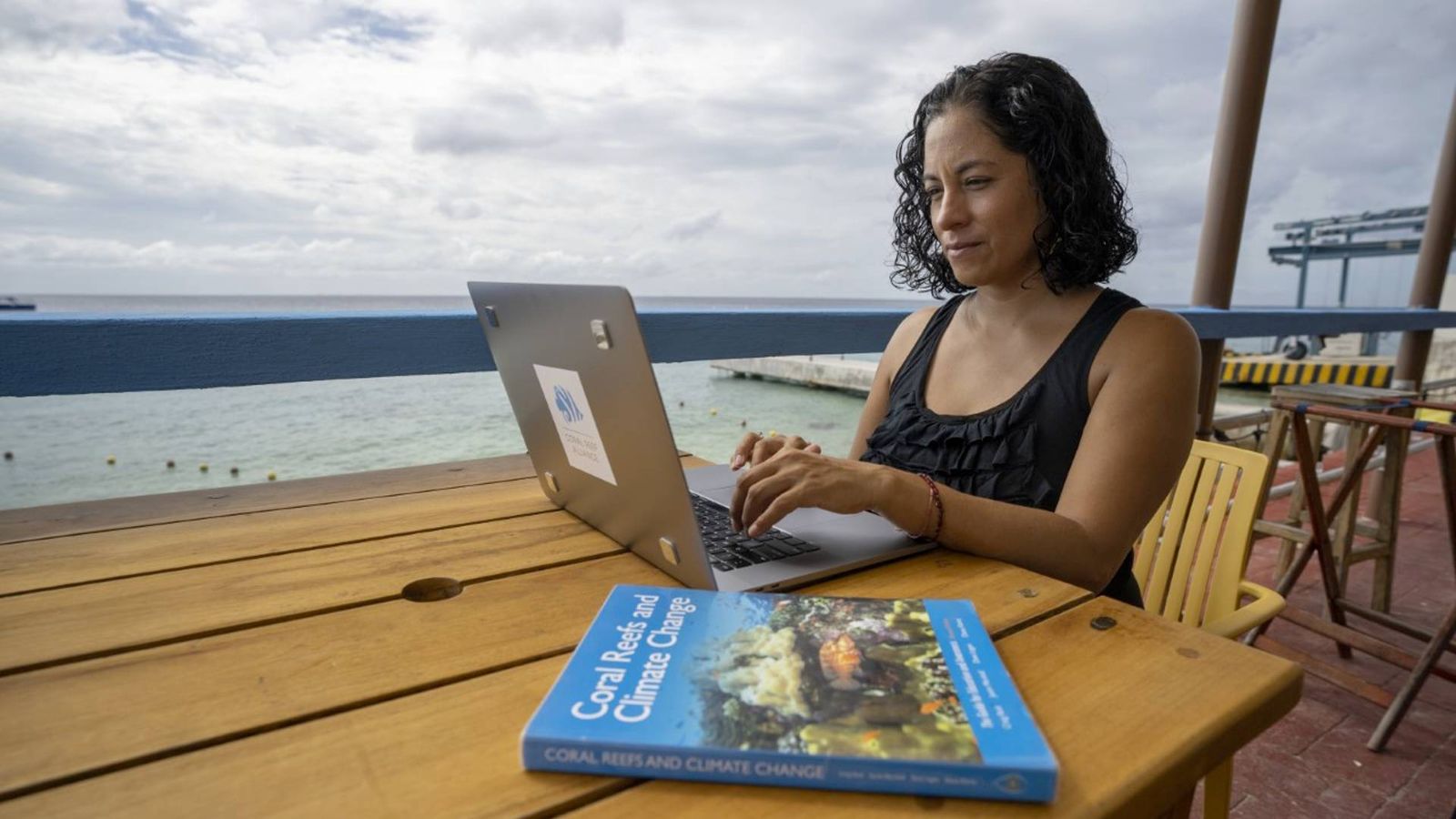 A woman working on a laptop with a coral reef textbook next to her.