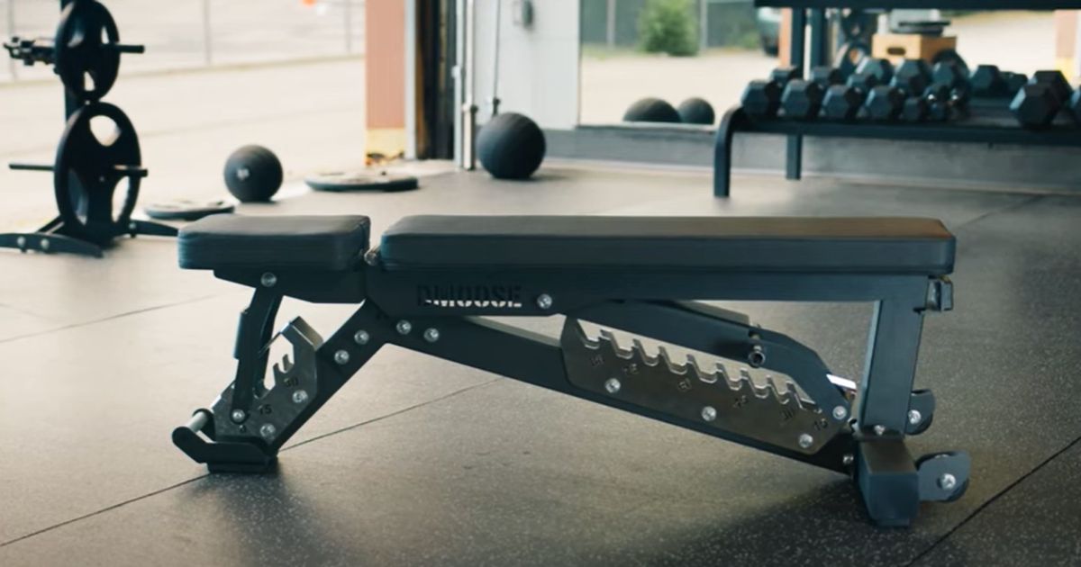 A black adjustable weight bench in a gym in front of a dumbbell rack and weight plates.