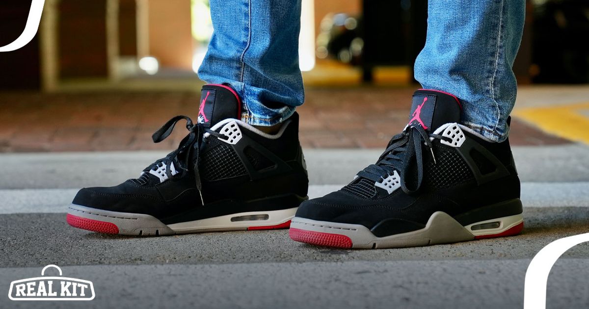 Someone in blue jeans wearing a pair of black pair of Jordan 4s featuring grey and red details and branding.