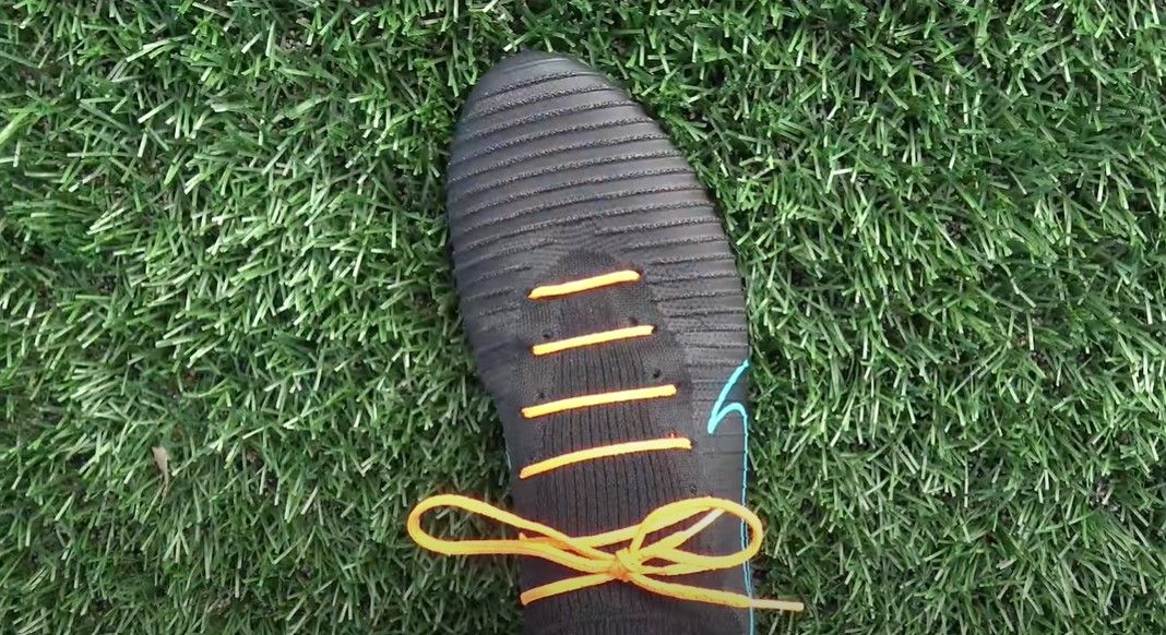 A black Nike football boot with an orange shoelace in straight bars across the middle.