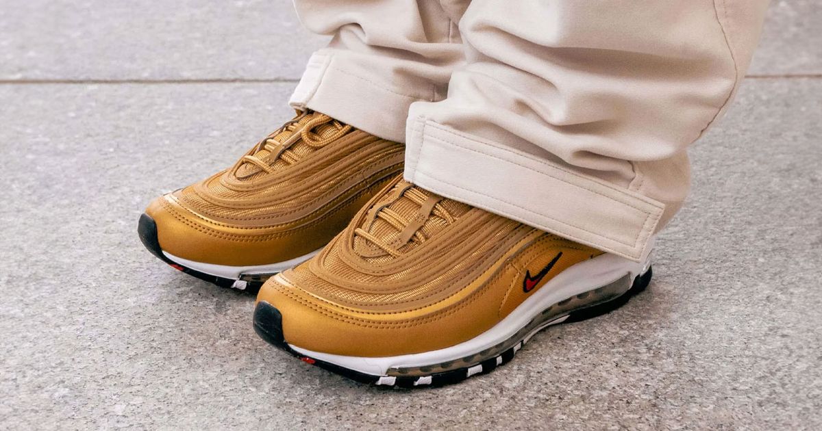 Someone in light cream trousers wearing a pair of gold Air Max sneakers with white midsoles and small red Swooshes on the sides.