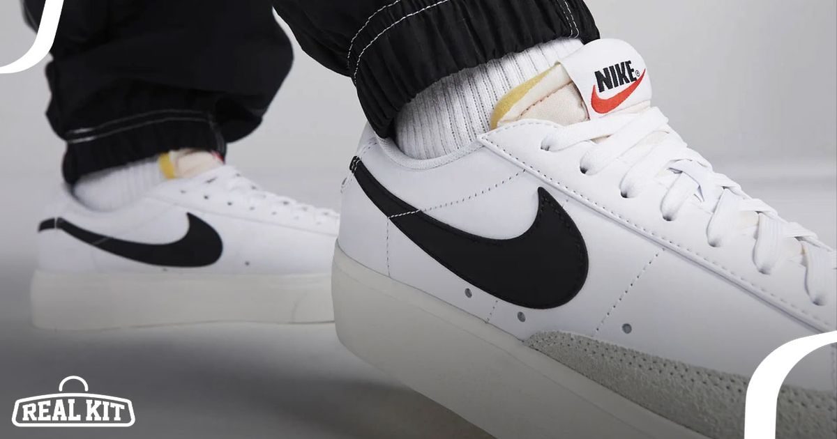 A close-up of someone in black trousers and white socks wearing a pair of white Nike Blazer Lows featuring black Swooshes down the sides.