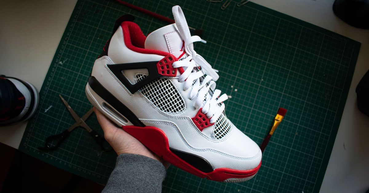 Someone in a grey top holding a white Jordan 4 featuring red and black trim and details.