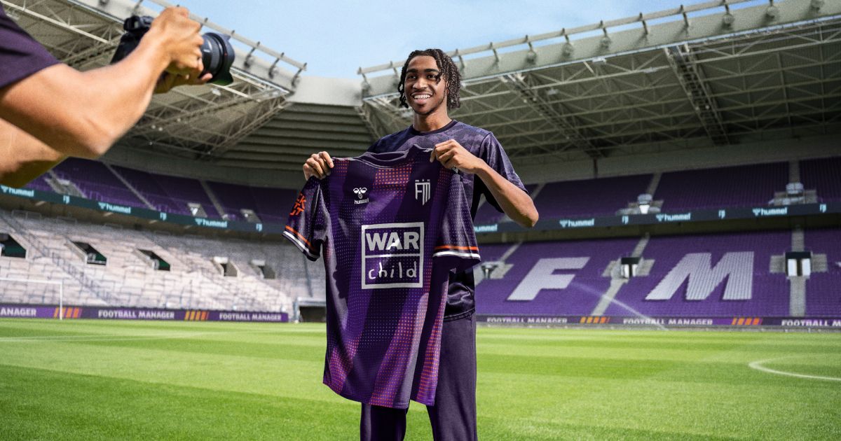 Someone standing on the pitch in a stadium holding a purple shirt while having their photo taken.