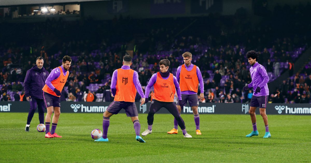 A team dressed in purple kit and orange bibs warming up playing football in a stadium in front of a manager in a dark purple coat.